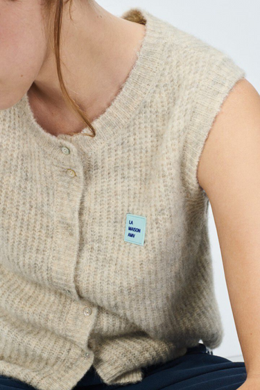 Model wearing the American Vintage beige sleeveless cardigan with logo in blue. Close front view